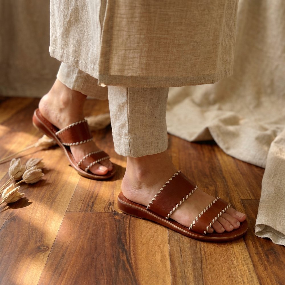 Handcrafted brown leather dual-strap slippers for women with stitched detailing on wooden background